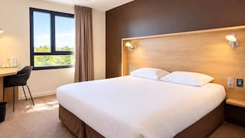 A hotel room with a large bed, a desk with a chair, a window with curtains, and a wall-mounted light fixture.