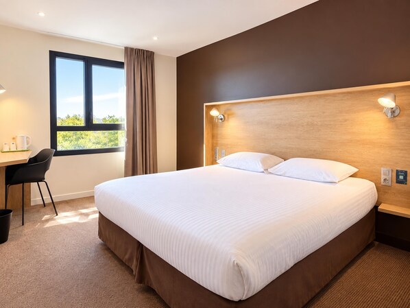 Une chambre d’hôtel avec un grand lit, un bureau avec une chaise, une fenêtre avec des rideaux et un luminaire fixé au mur.
