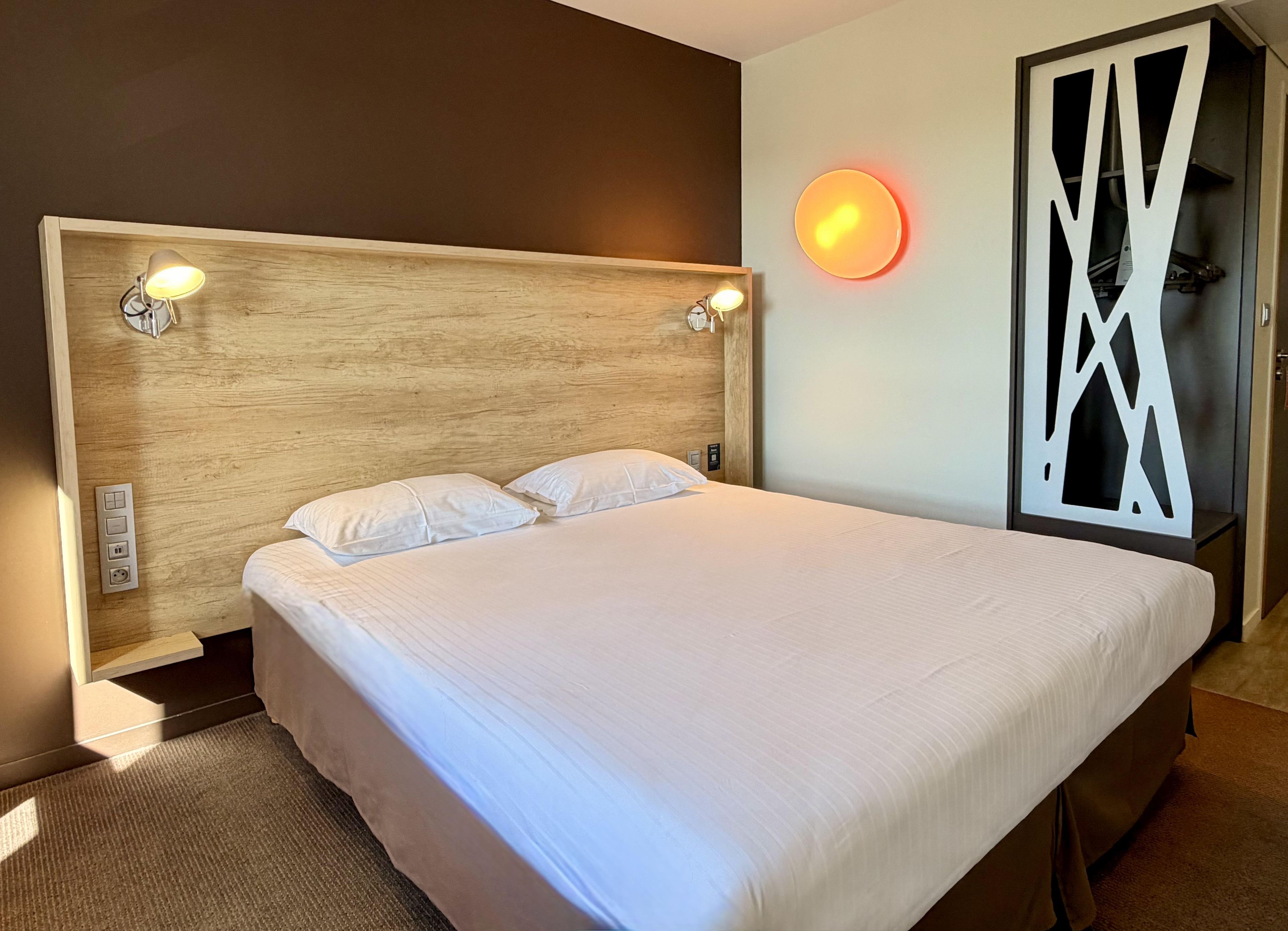 Une chambre d’hôtel moderne avec un grand lit, des lampes de chevet, un luminaire fixé au mur et un panneau décoratif.