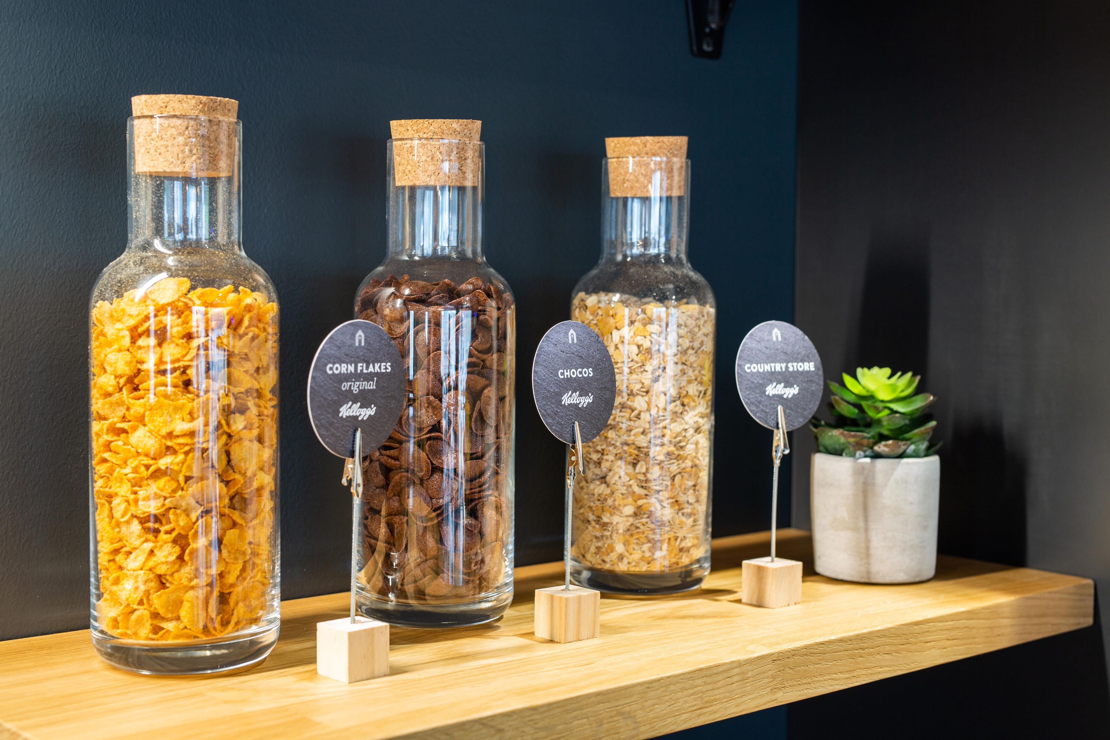 Four glass jars with labels on a wooden shelf, each containing different types of cereal.