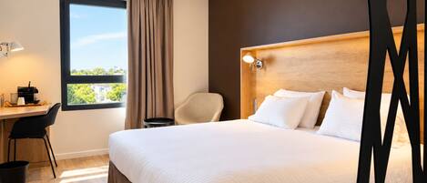A hotel room with a large bed, a desk with a chair, a view of the outdoors, and a wooden headboard.