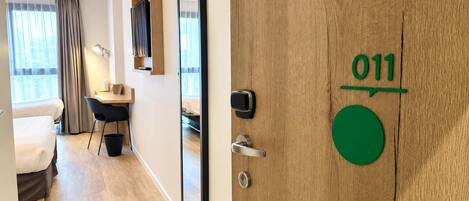 A hotel room with a wooden door marked '011', a desk with a chair, a bed, and a window with curtains.