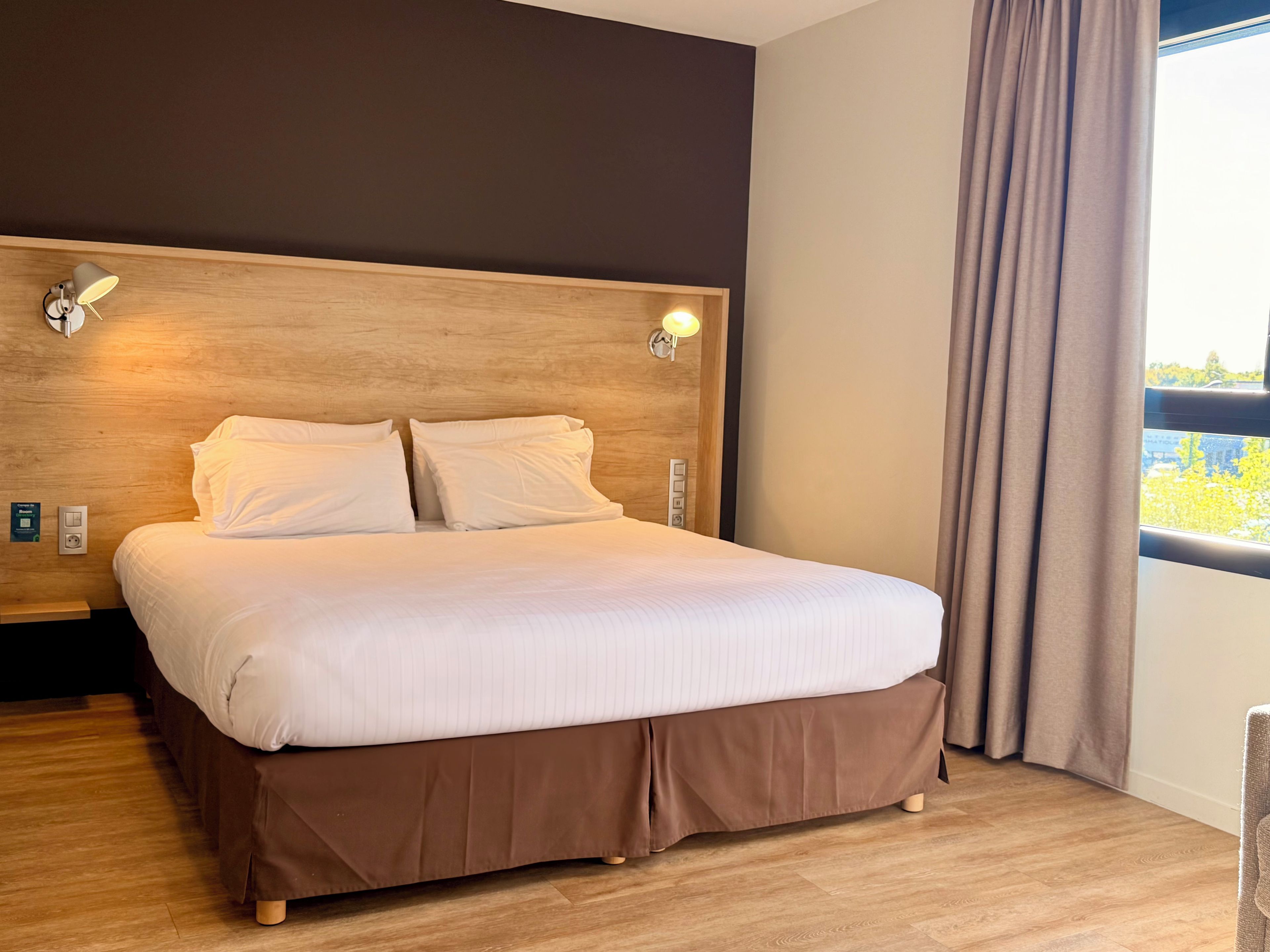 Cama bem arrumada com cabeceira de madeira, duas luminárias fixadas na parede e uma janela com cortinas.