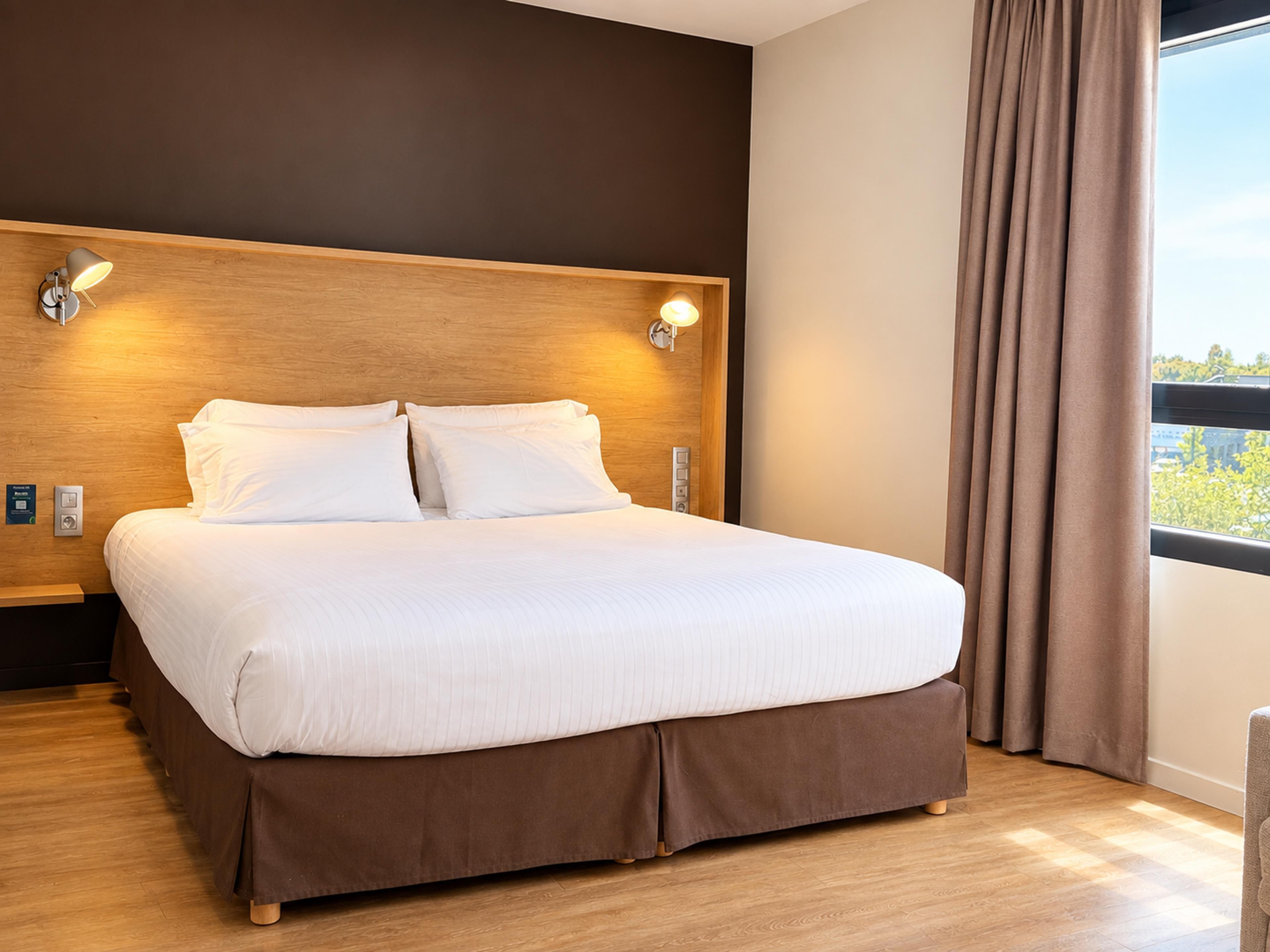 Una cama bien tendida con cabecera de madera, dos lámparas de noche y vistas al exterior a través de una ventana con cortinas.