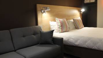 Una habitación de hotel moderna con un sofá gris oscuro, una cama grande con almohadillas estampadas y dos lámparas de lectura fijadas a la pared.