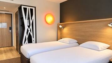 Une chambre d’hôtel avec deux lits, une tête de lit en bois, une lampe fixée au mur et une porte ornée d’un panneau décoratif.