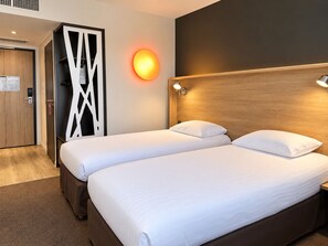 Une chambre d’hôtel avec deux lits, une tête de lit en bois, une lampe fixée au mur et une porte ornée d’un panneau décoratif.