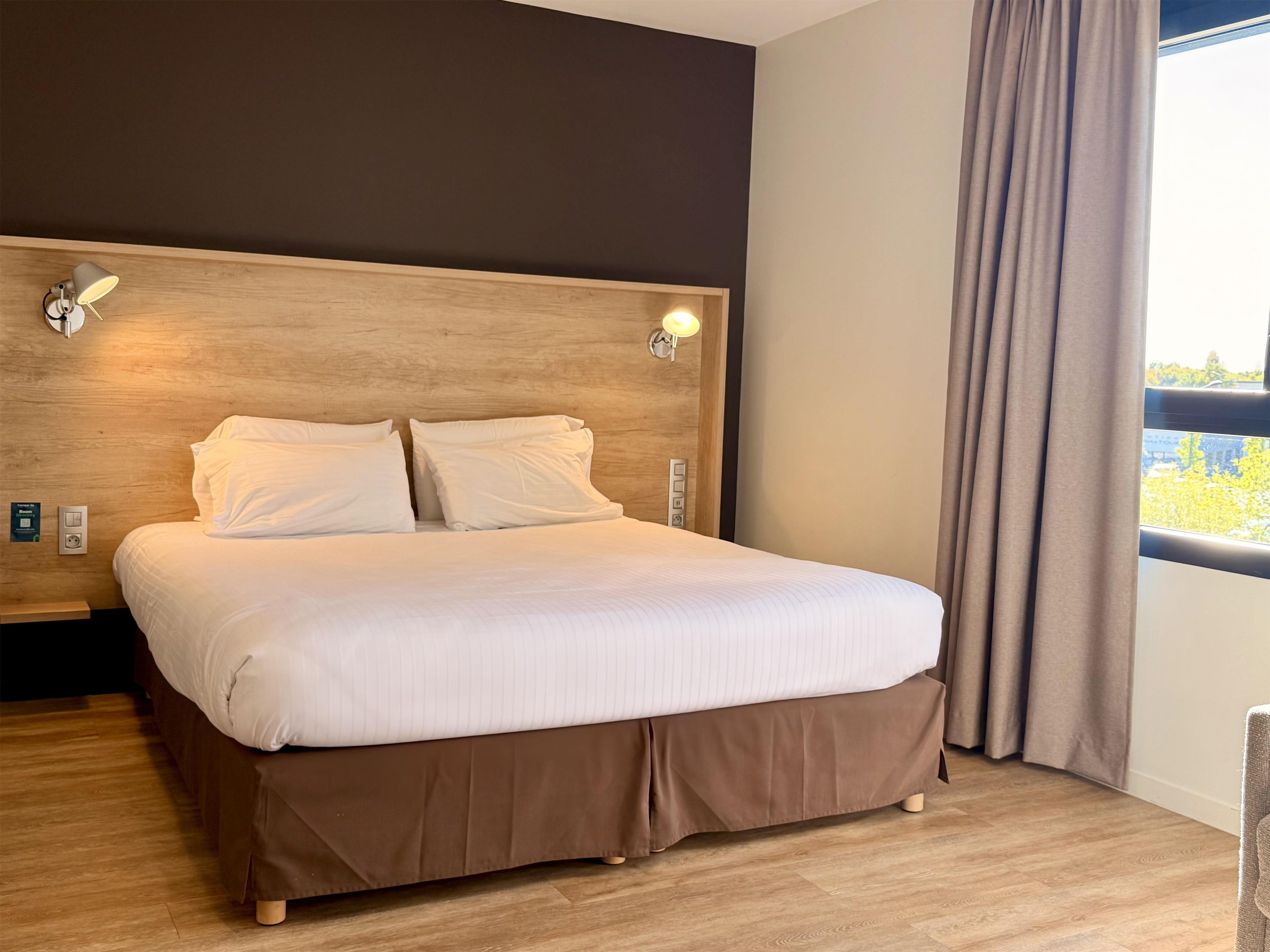 A neatly made bed with a wooden headboard, two wall-mounted lights, and a window with curtains.