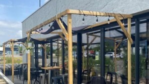 Een terras buiten met houten pergola's en tafels, omringd door glazen wanden.