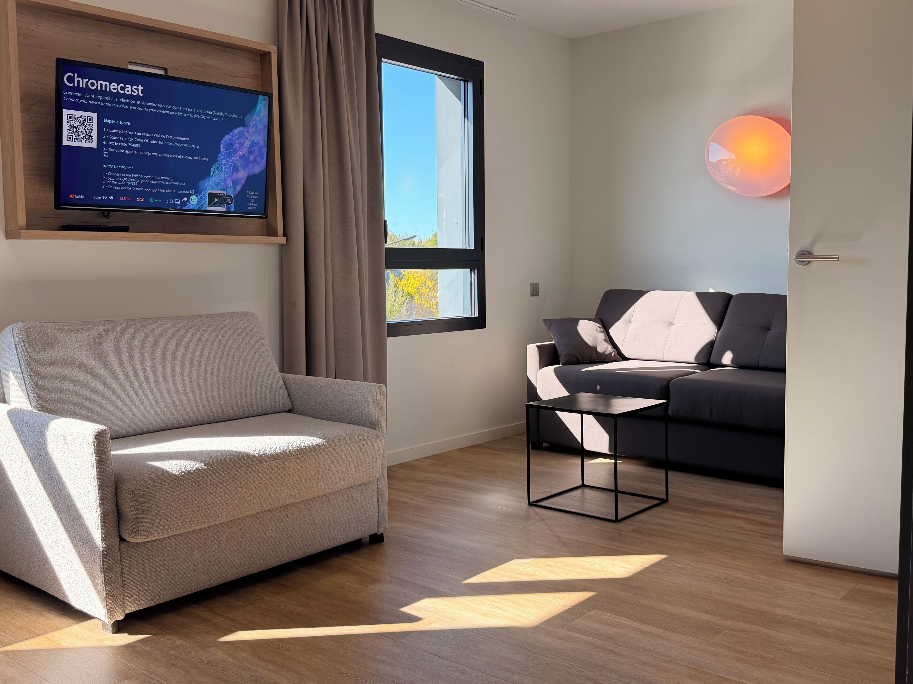 A modern living room with a grey armchair, a black and white sofa, a small black table, and a wall-mounted TV displaying a Chromecast setup guide.