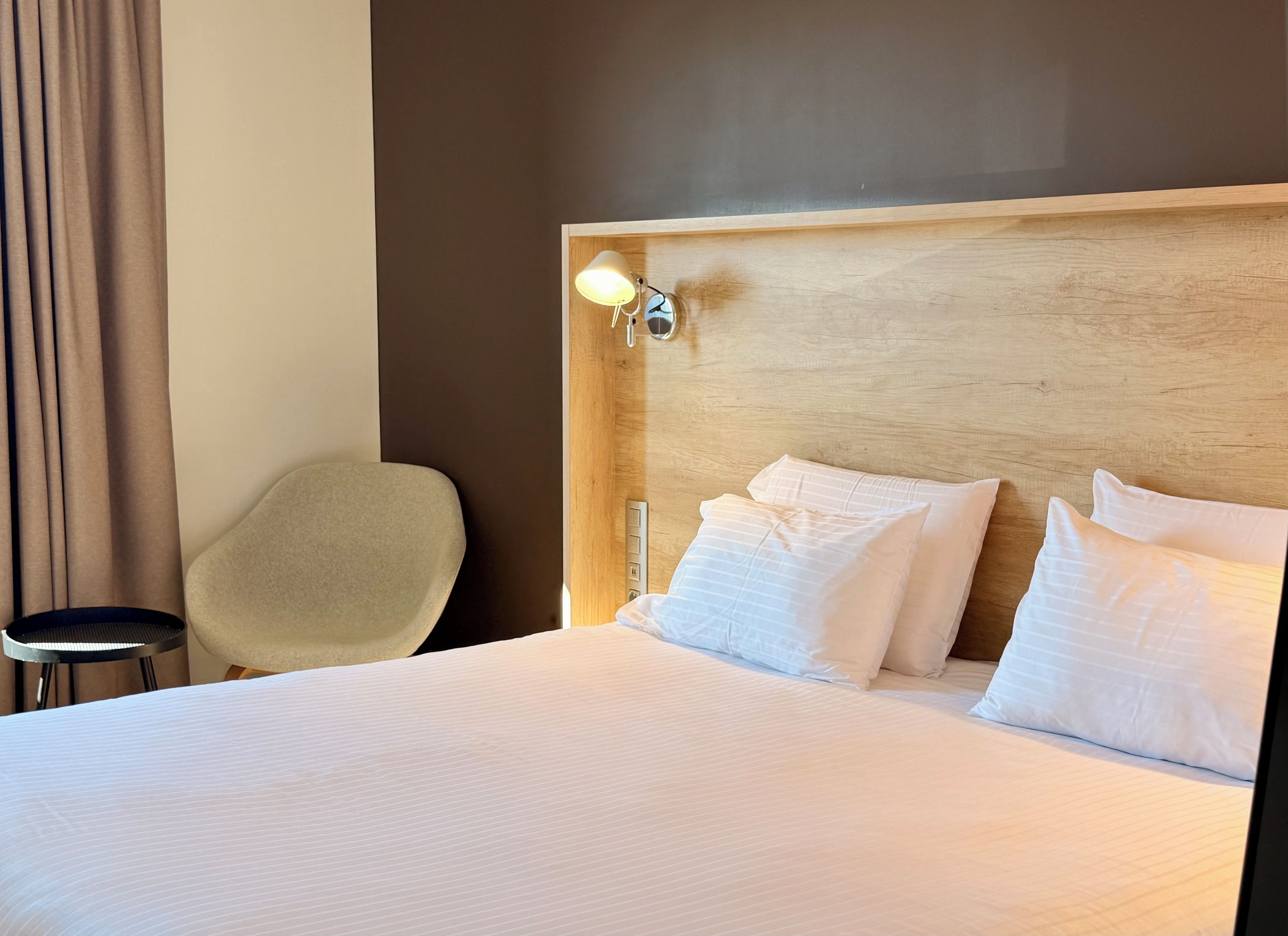Una habitación de hotel con una cama, una silla, una mesita de noche y una lámpara de pared.