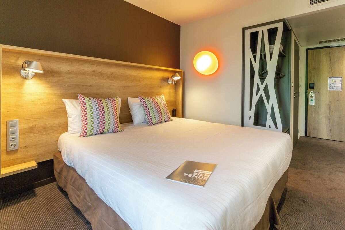 A hotel room with a large bed, two pillows, a bedside table, a lamp, and a wall-mounted light.
