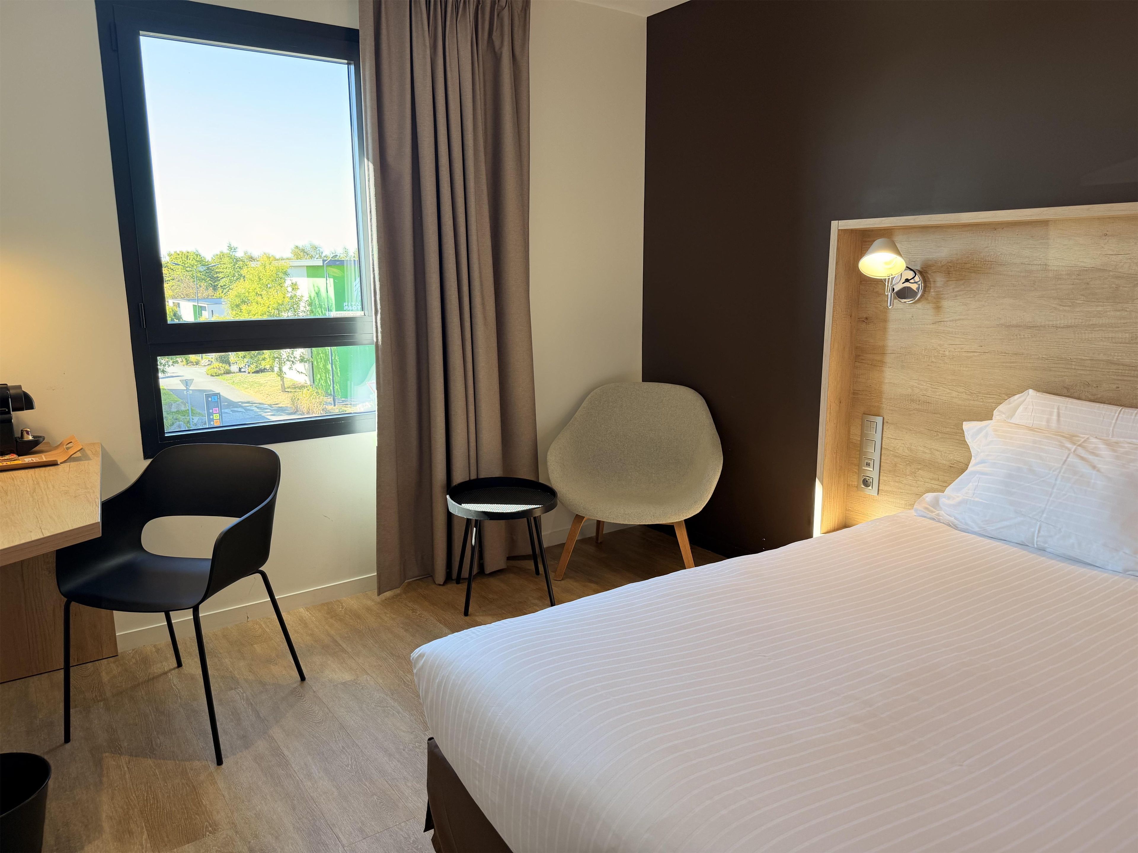 A hotel room with a bed, a desk, two chairs, a window with curtains, and a view of the outside.