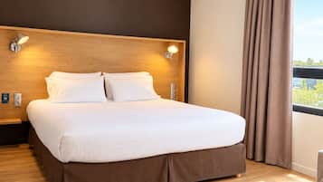 A hotel room with a large bed, wooden headboard, two wall-mounted lights, a window with curtains, and a wooden floor.