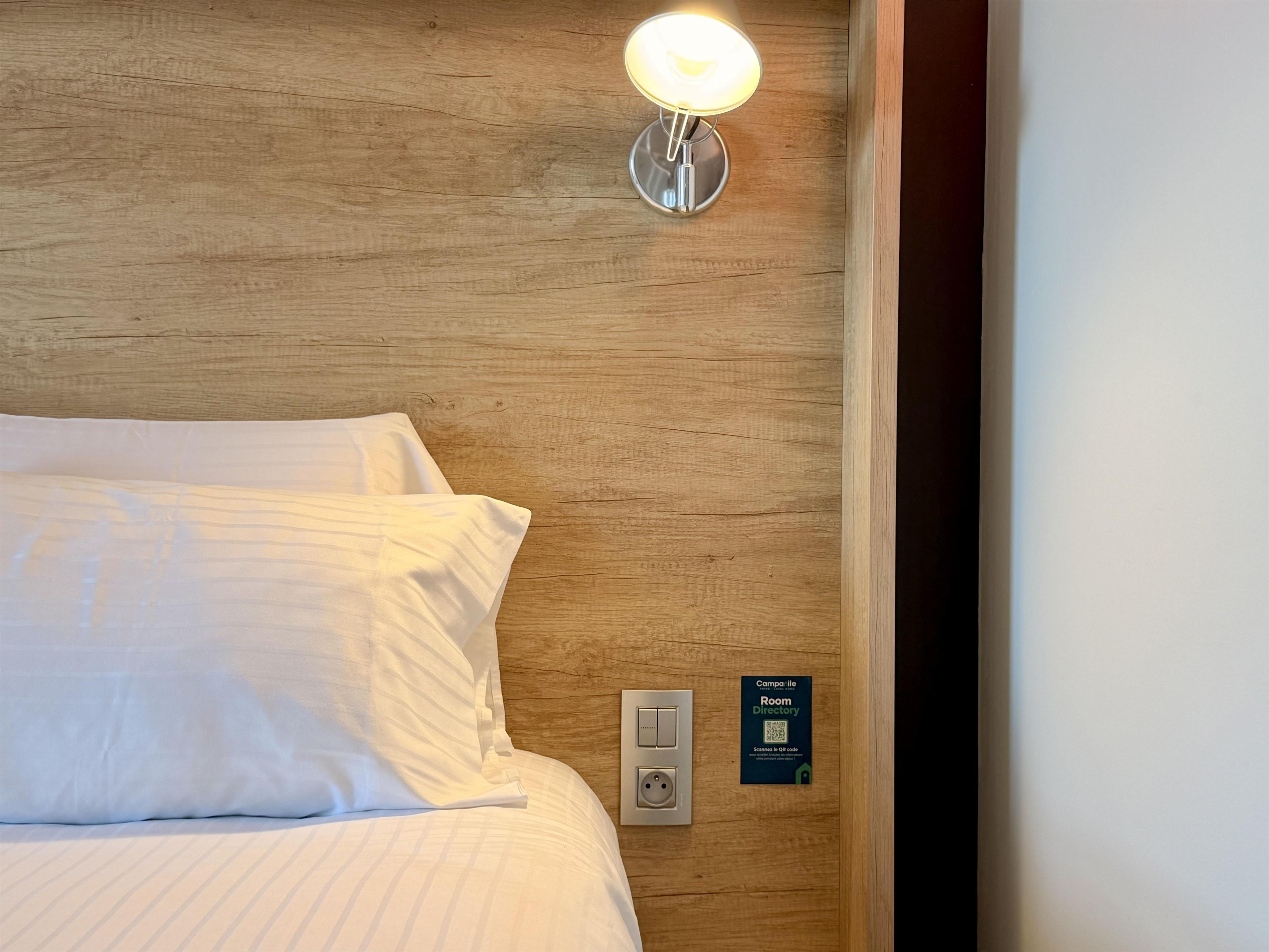 Una cama bien tendida con sábanas blancas, un aplique en la pared y un código QR para la guía de la habitación.