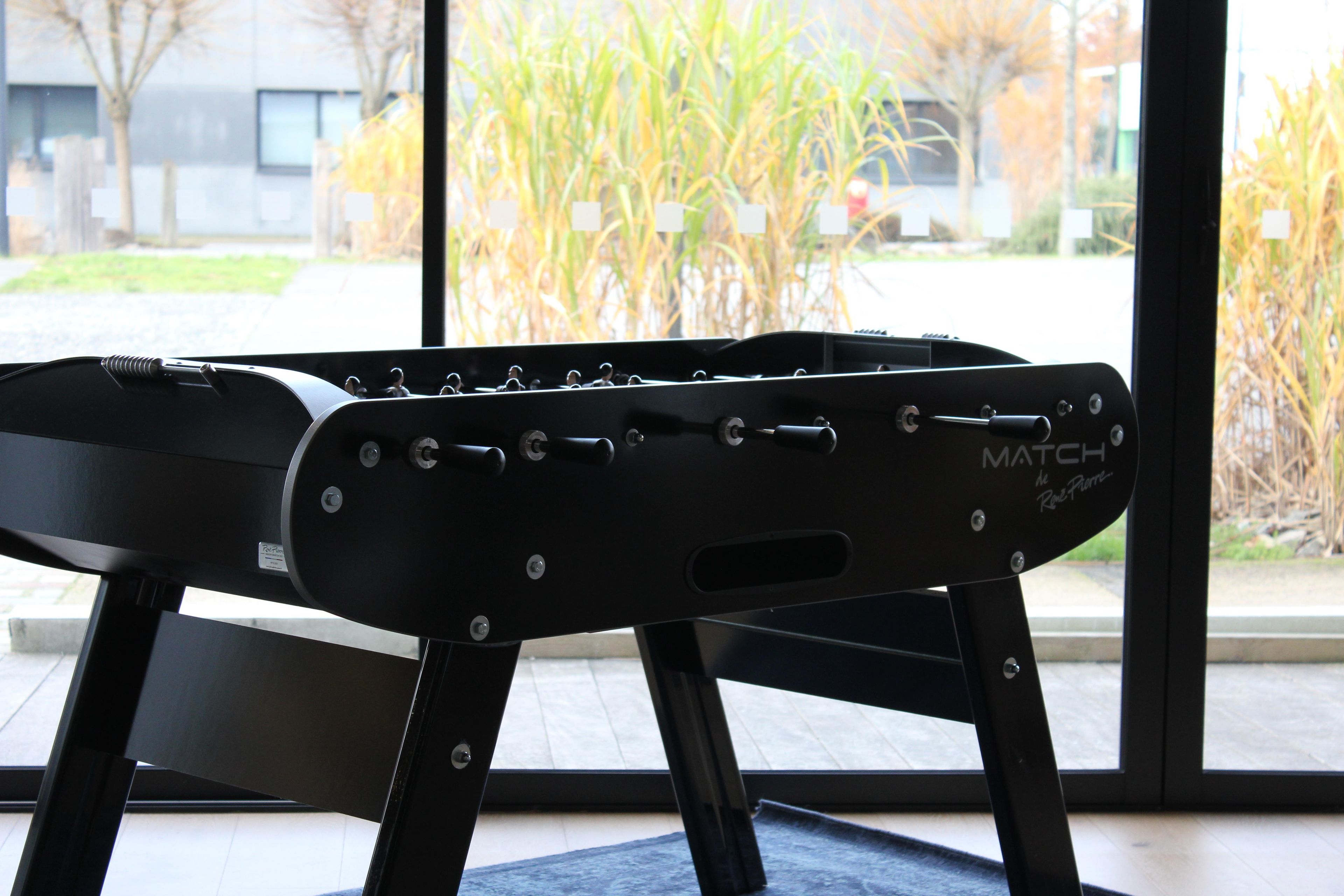 Una máquina de ejercicio negra con la marca 'MATCH de Rene Pierre', ubicada en un interior con vista a un edificio y follaje exterior.