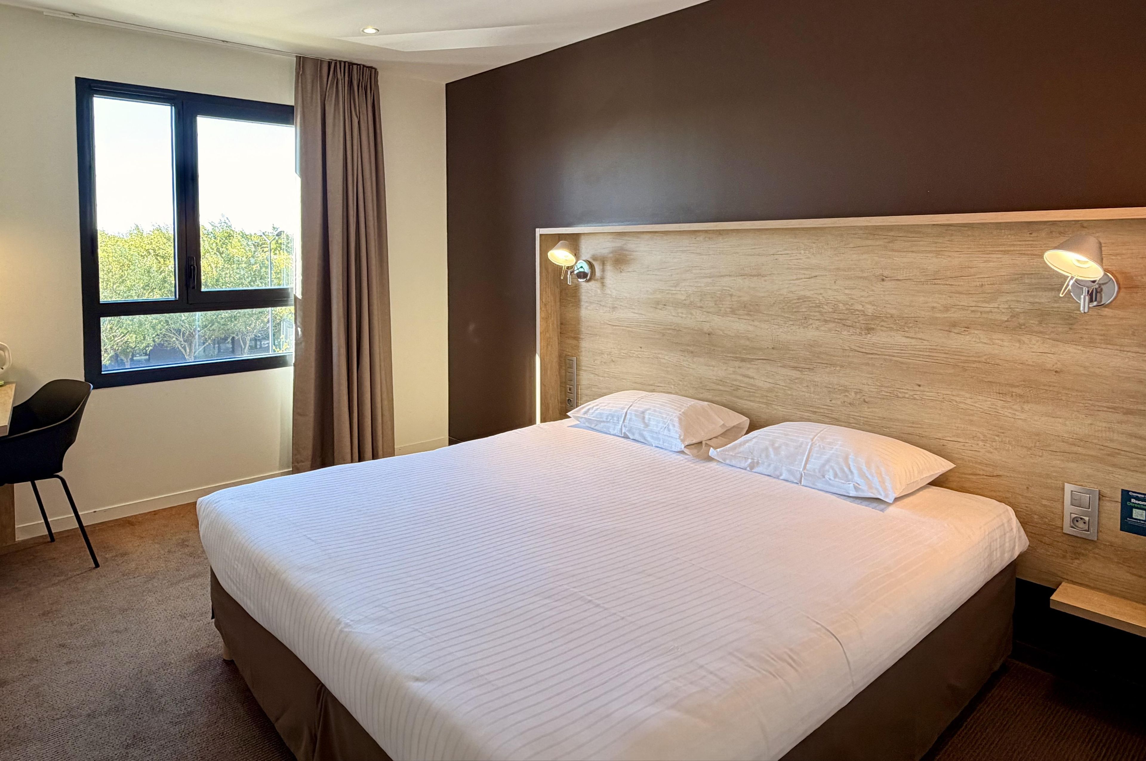 A hotel room with a large bed, a desk, a chair, and a window with curtains.