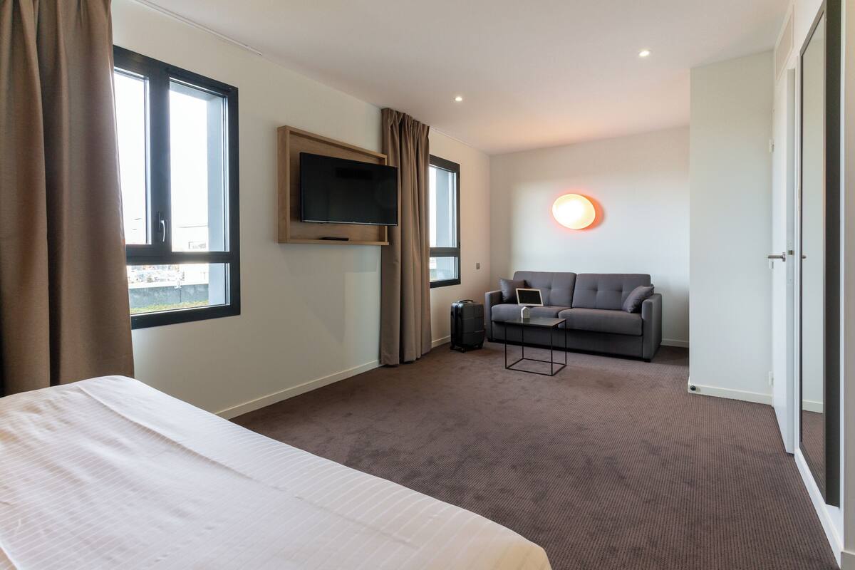 A hotel room with a bed, a sofa, a TV, and a window.