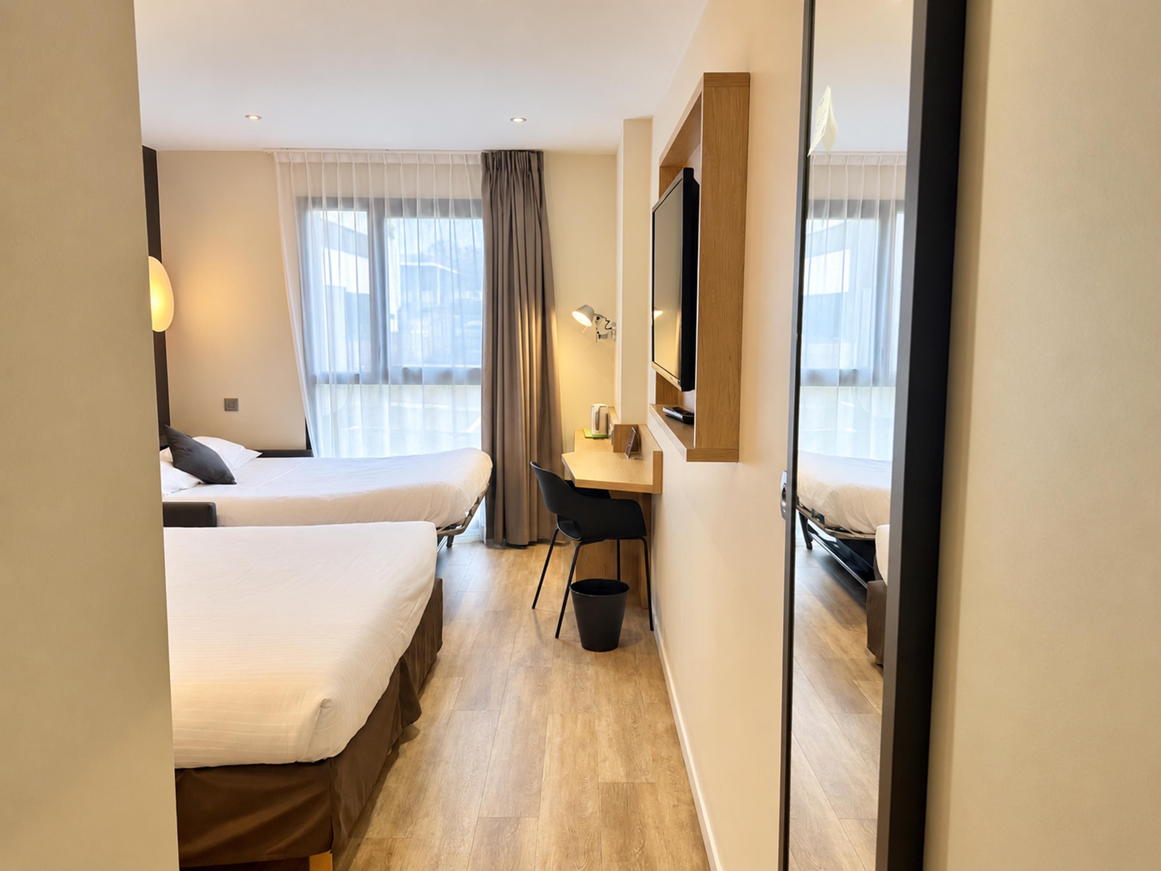 Habitación de hotel con dos camas, un escritorio, una silla, un televisor y una ventana con cortinas.