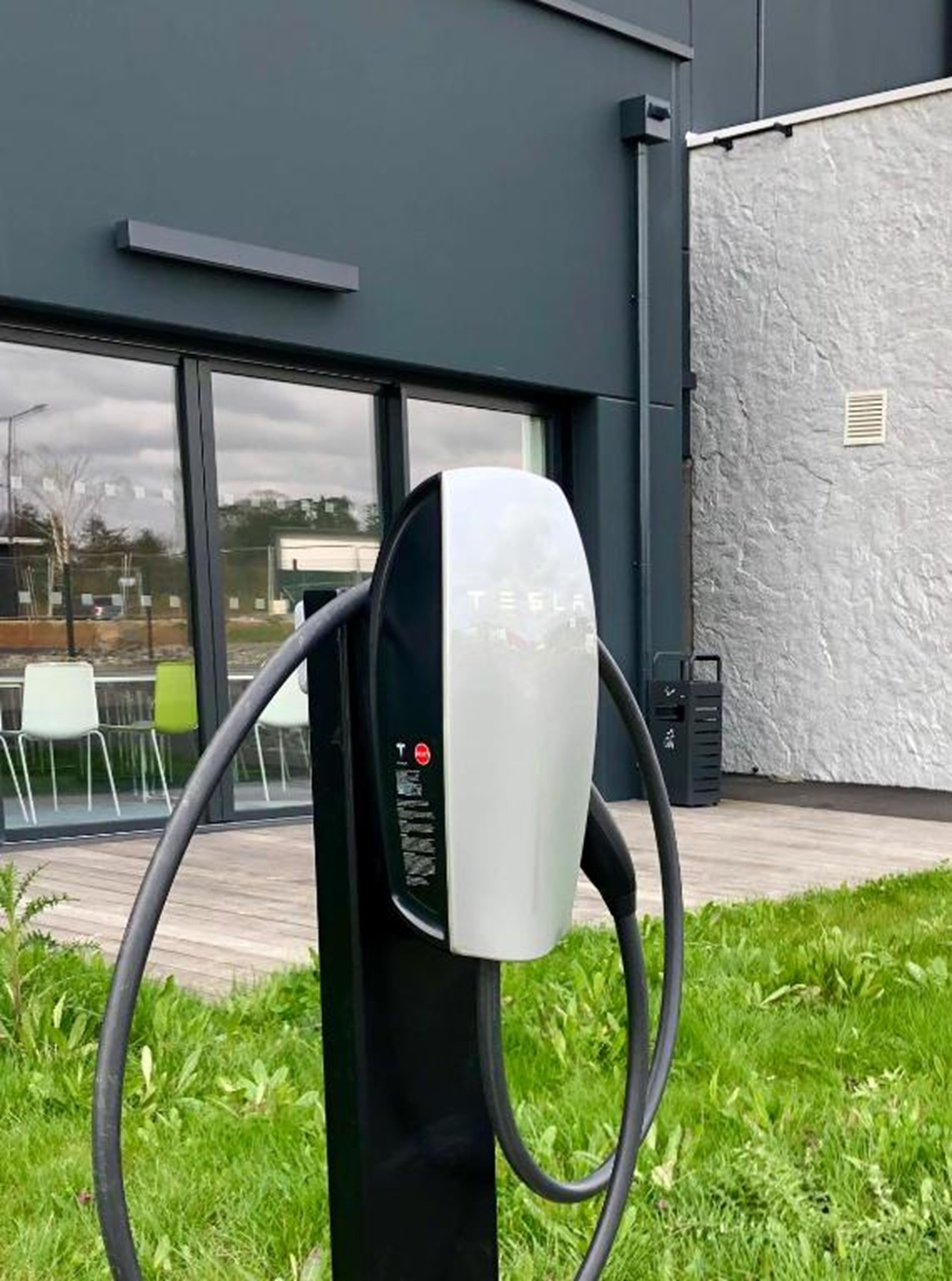 Een Tesla elektrische laadpaal met een zwart-wit ontwerp, buiten gelegen voor een modern gebouw met grote ramen en een gazon.