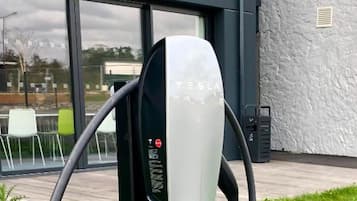 Une borne de recharge pour véhicules électriques Tesla, de couleur noire et blanche, située à l’extérieur, devant un bâtiment moderne doté de grandes fenêtres et bordée d’une zone verdoyante.