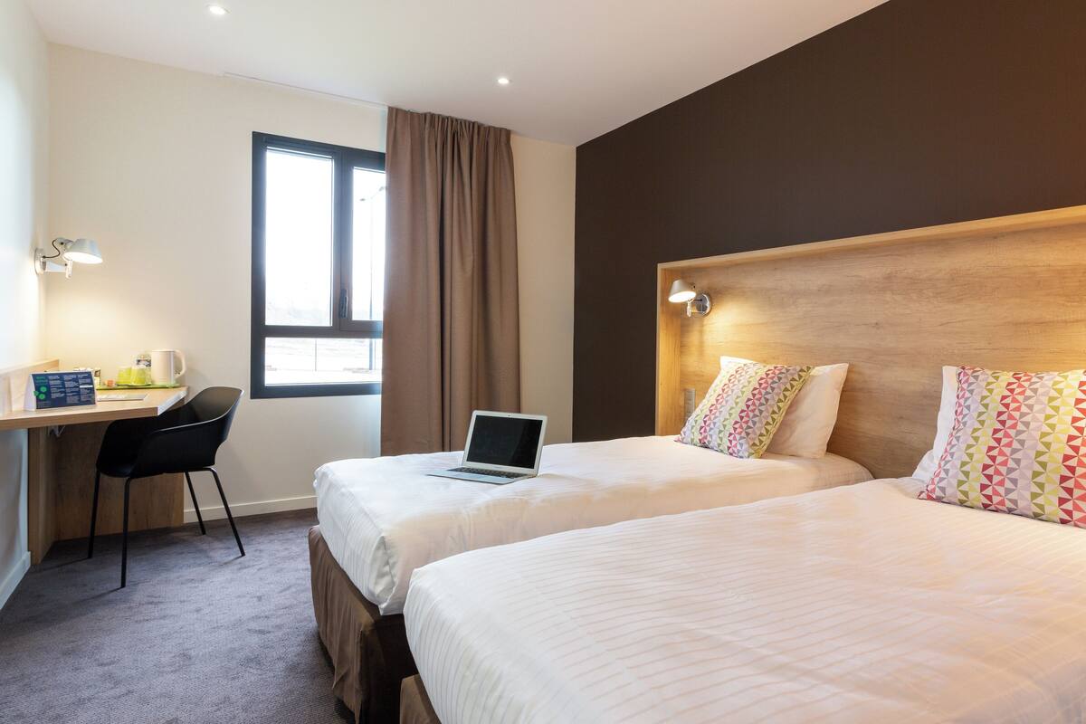 A hotel room with two beds, a desk with a laptop, a chair, and a window with curtains.