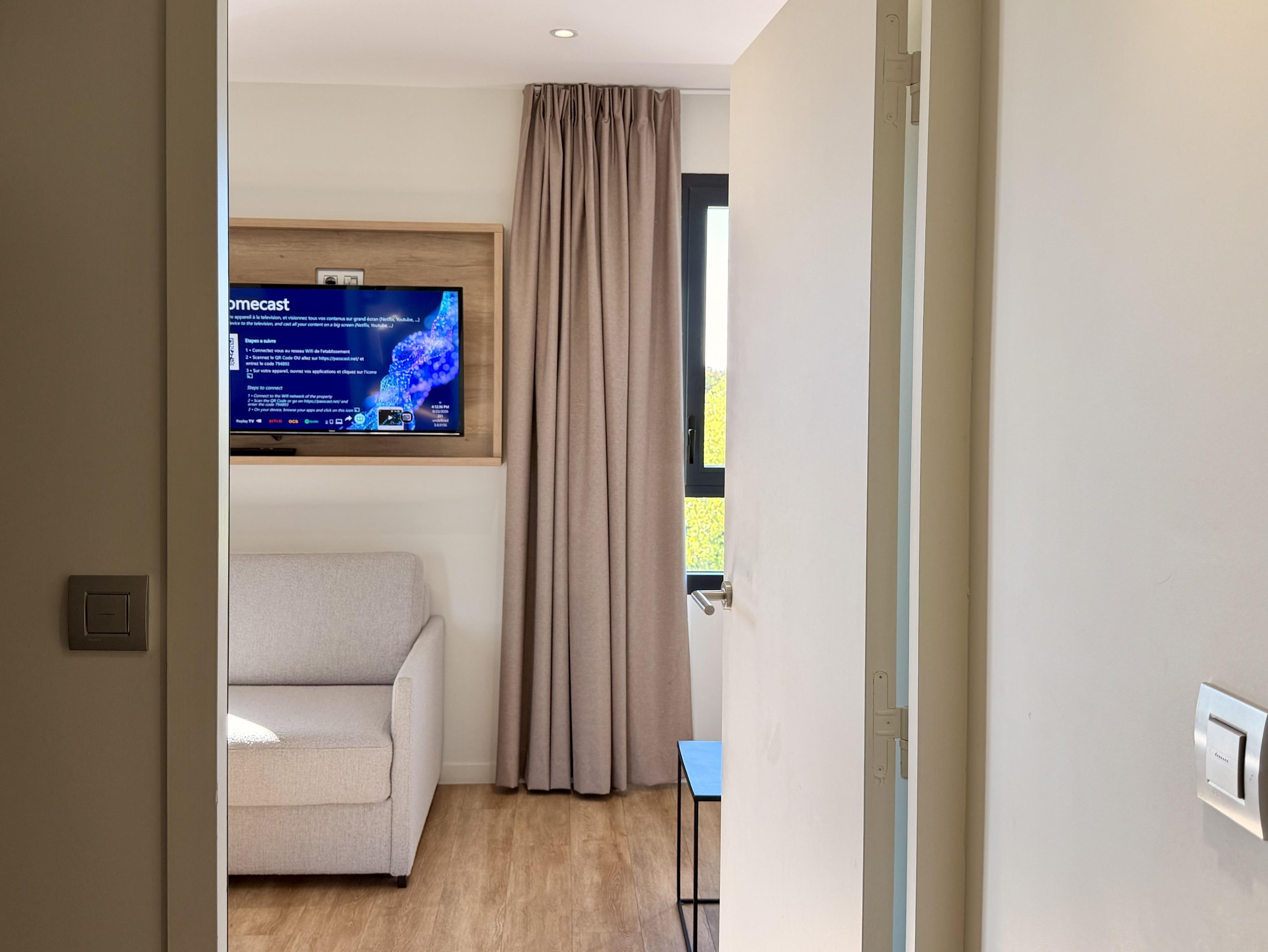 Et moderne værelse med et fladskærms-TV ophængt på væggen, en beige sofa, et lille bord og et vindue med gardiner.