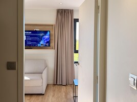 Una habitación moderna con una televisión de pantalla plana montada en la pared, un sofá color beige, una mesita y una ventana con cortinas.