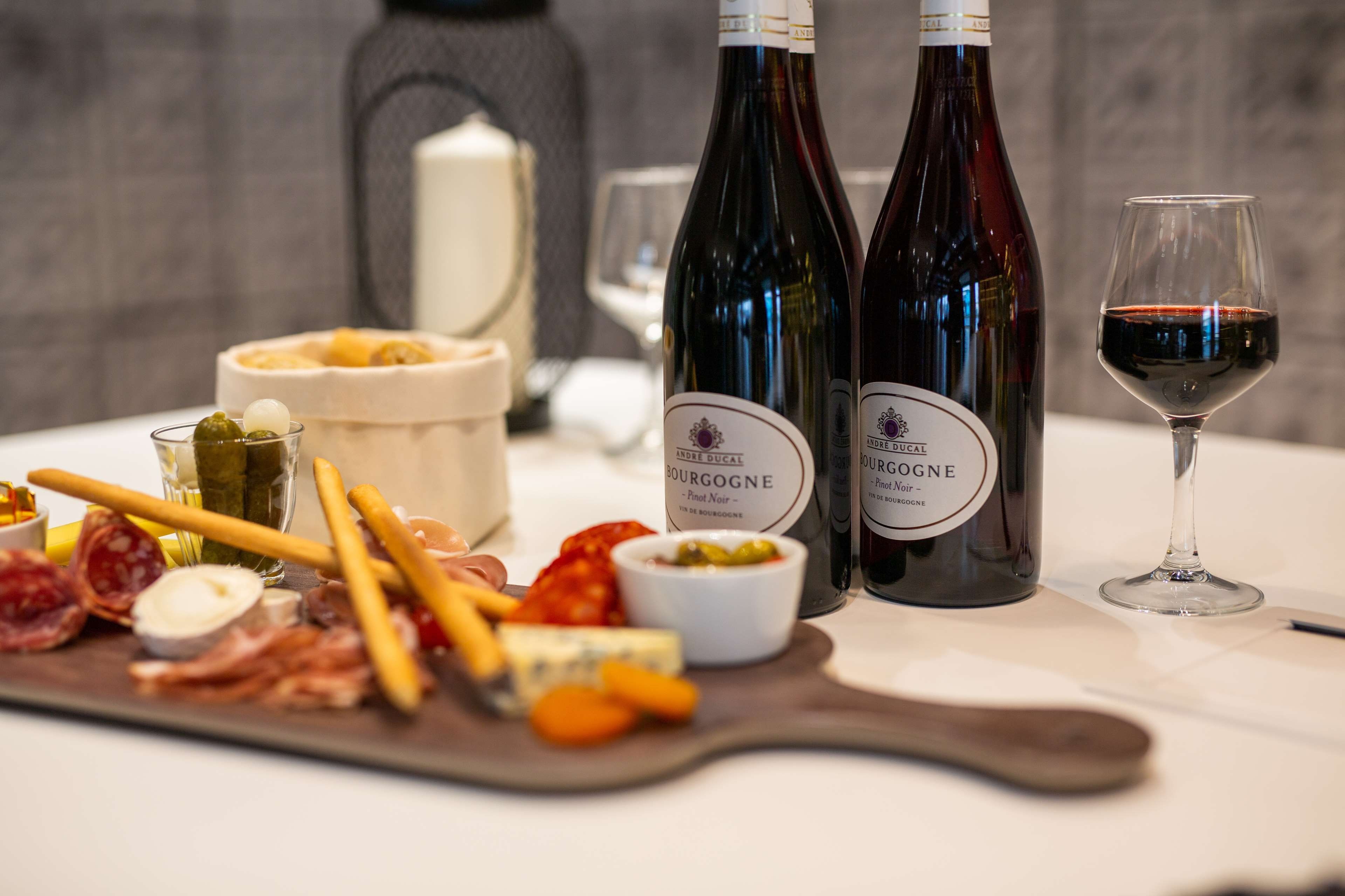 A table with a variety of cheeses, cured meats, and pickles, accompanied by two bottles of wine and a glass of red wine.