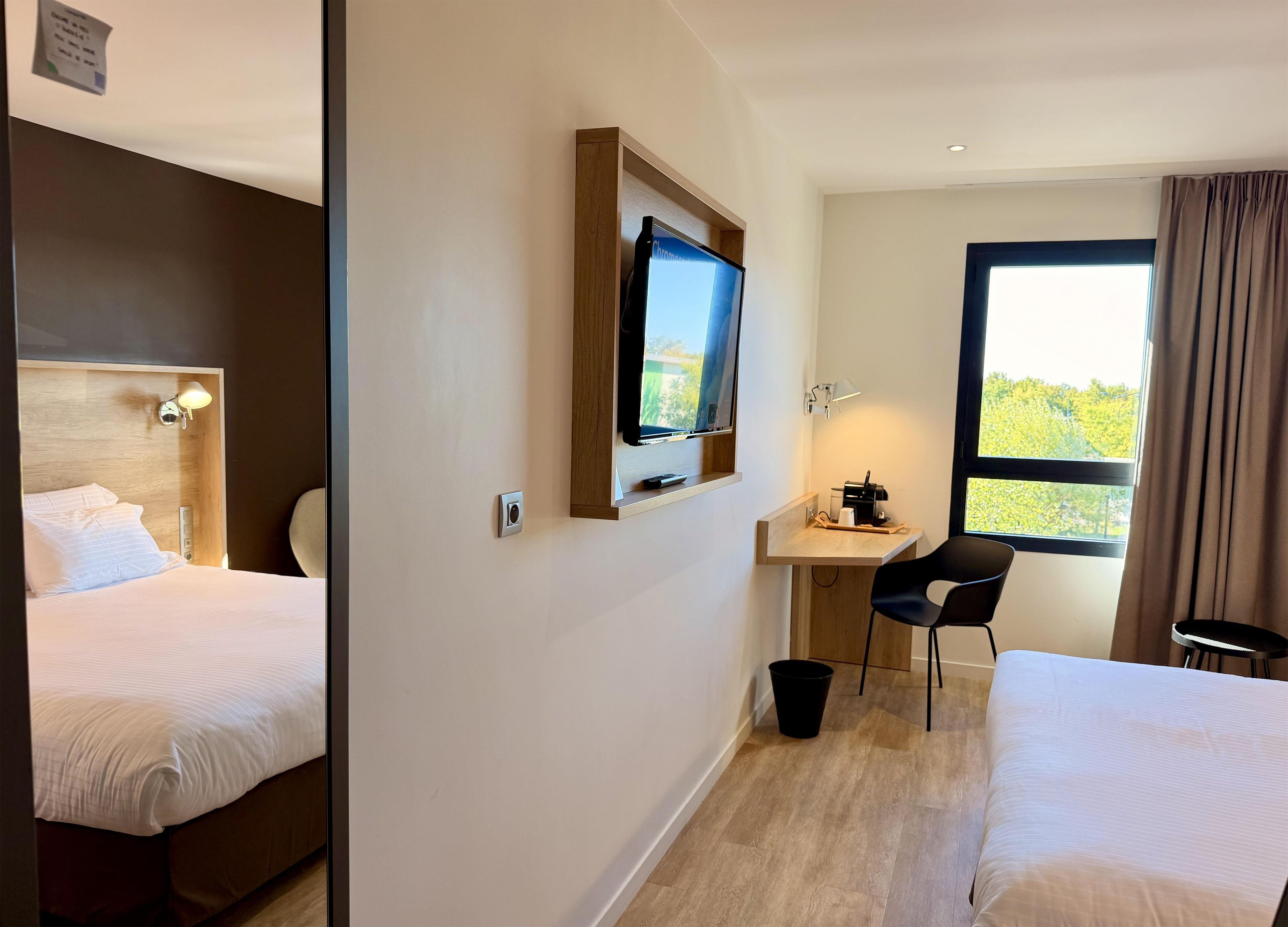 A hotel room with two beds, a desk with a chair, a television, and a window with curtains.
