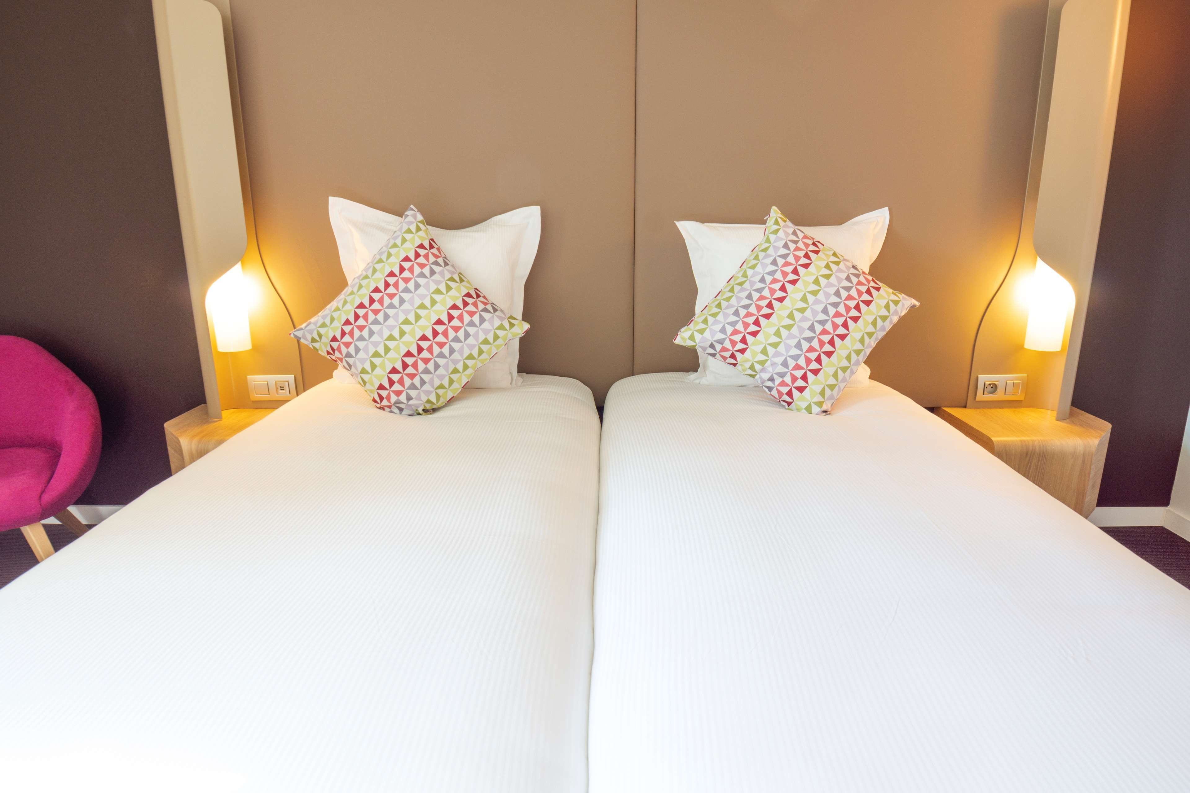 A hotel room with two beds, each with a white bedspread and a colorful geometric-patterned pillow. There are bedside lamps on either side of the beds.