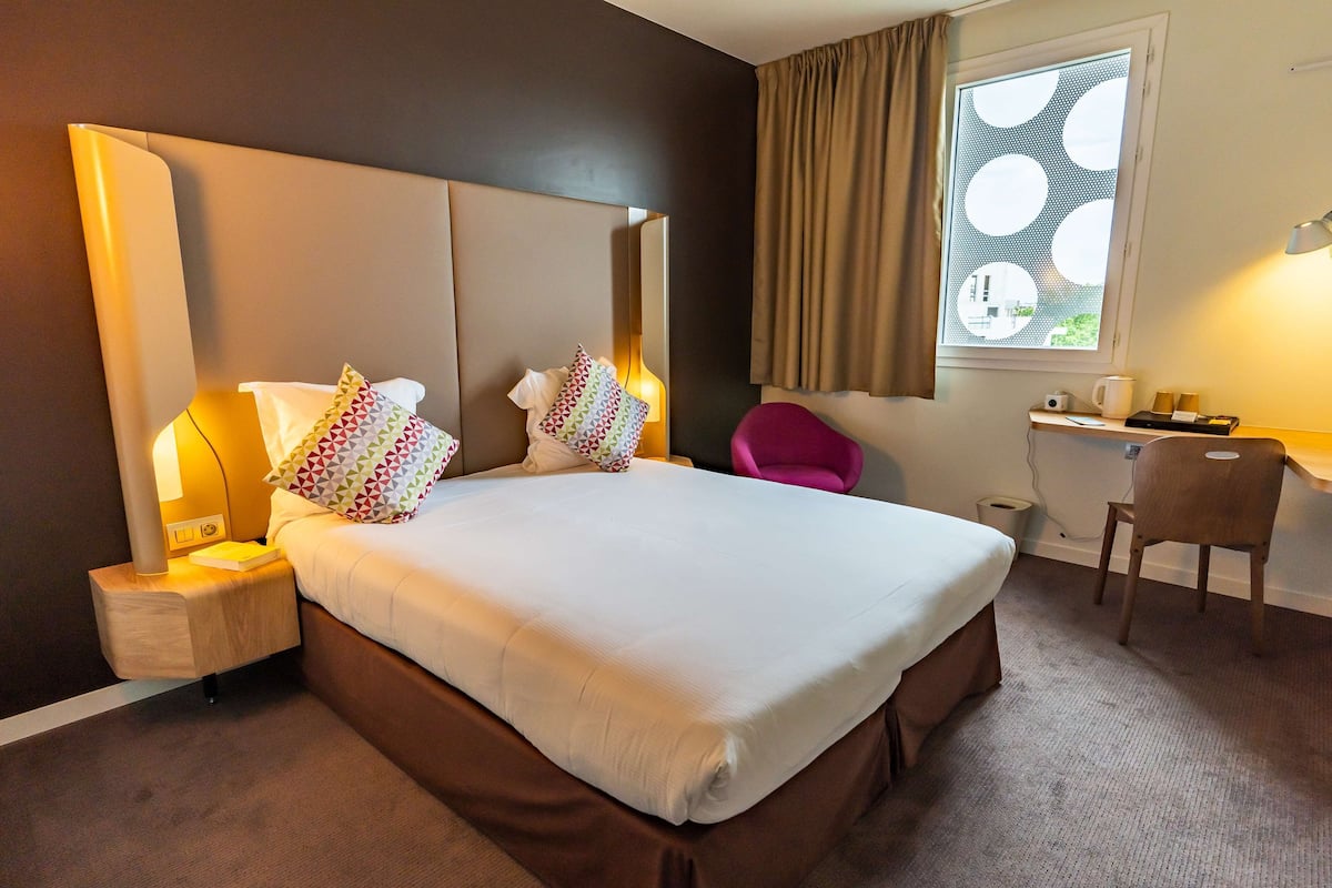 Une chambre d’hôtel avec un grand lit, des tables de chevet, un bureau, une chaise et une fenêtre avec un rideau.