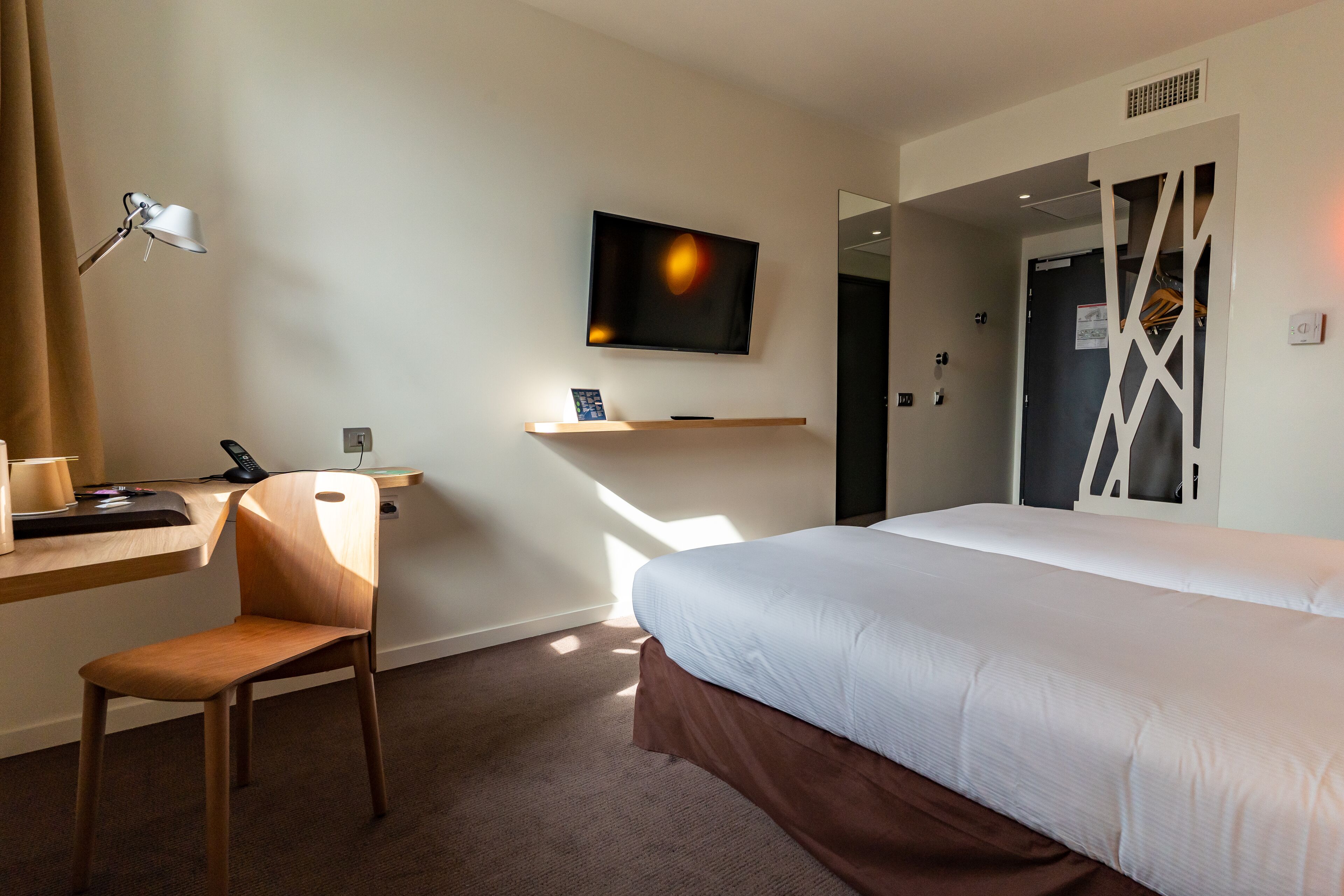 Habitación de hotel con cama, escritorio con silla, televisor montado en la pared y ventana que deja entrar luz natural.
