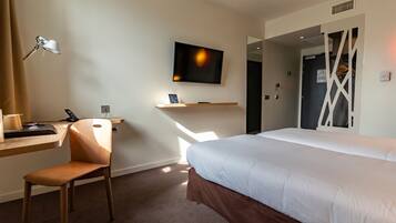 Habitación de hotel con cama, escritorio con silla, televisor montado en la pared y ventana que deja entrar luz natural.