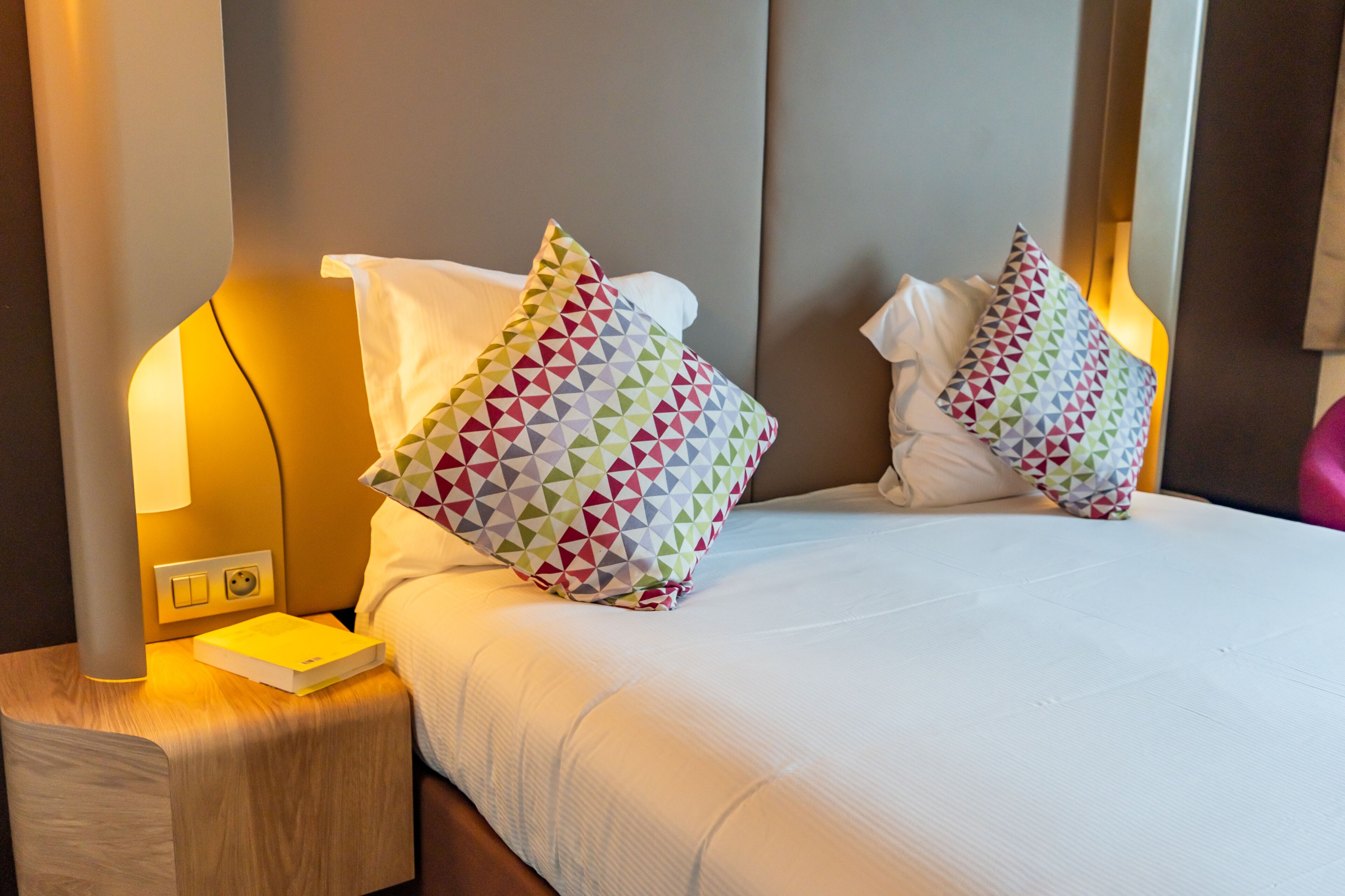Un lit bien fait, avec du linge de lit blanc et des coussins aux motifs géométriques et aux couleurs vives.