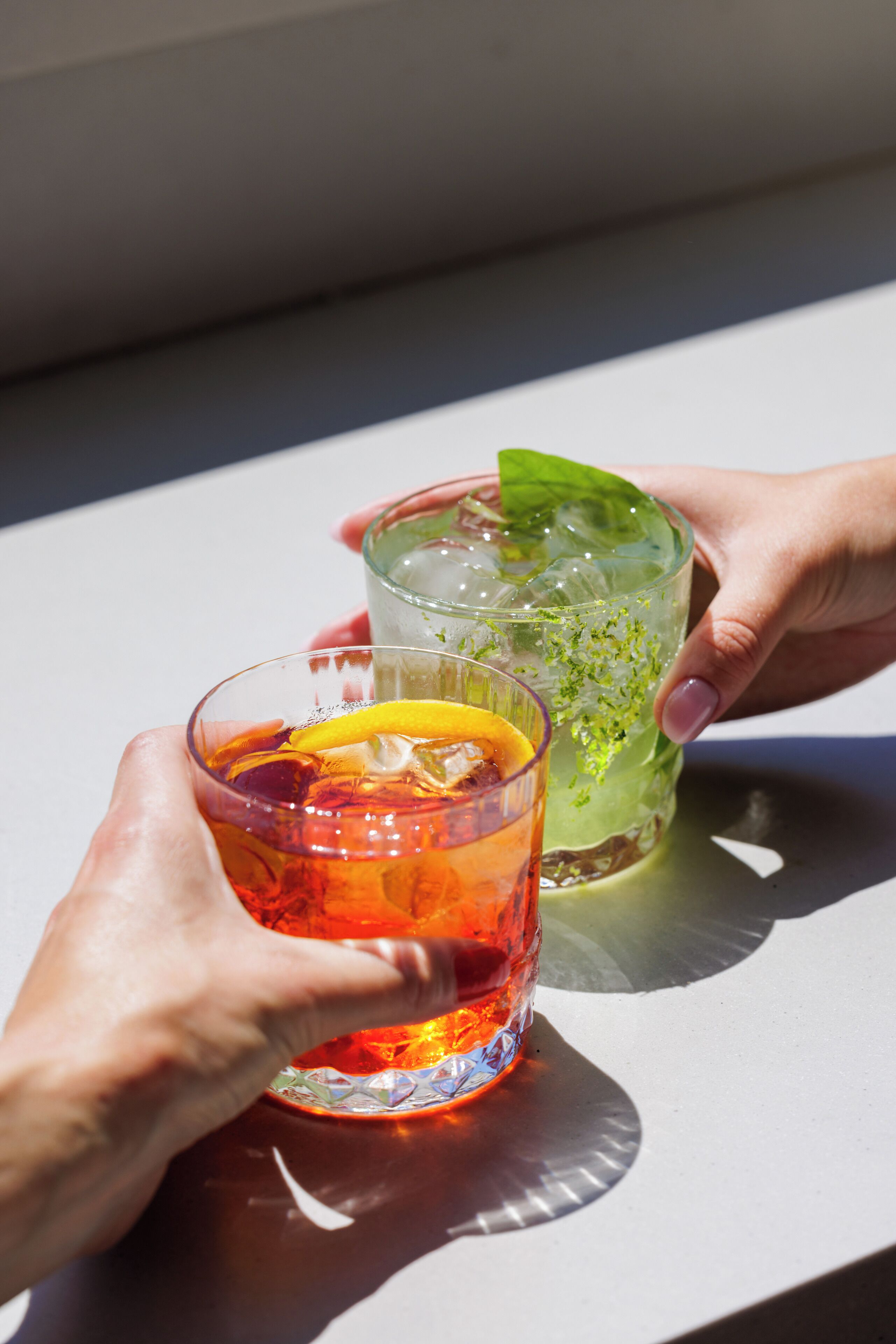 Two hands holding glasses of iced drinks with ice cubes and garnishes.