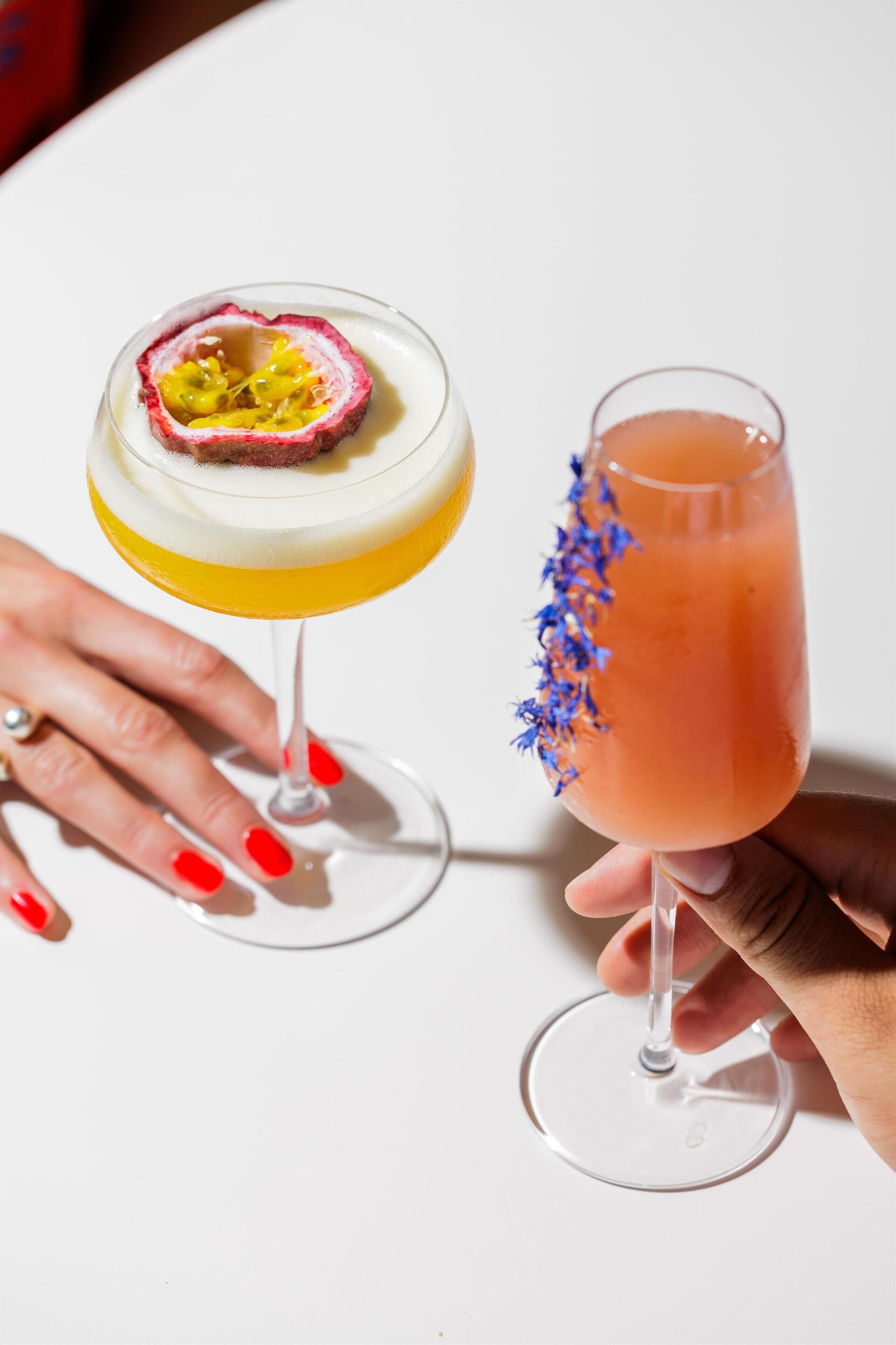 Two cocktails in wine glasses, one with a passion fruit slice and the other with a blue floral garnish.