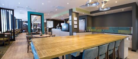 Un restaurante moderno con una gran mesa de madera, taburetes de bar, iluminación colgante y una zona de cocina al fondo.