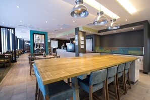 A modern restaurant with a large wooden table, bar stools, pendant lighting, and a kitchen area in the background. - Premier Inn Manchester Salford Media City (Salford)