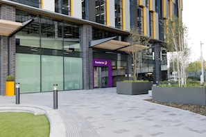 A modern hotel entrance with a glass facade, a prominent purple sign, and a landscaped area with planters. - Premier Inn Manchester Salford Media City (Salford)