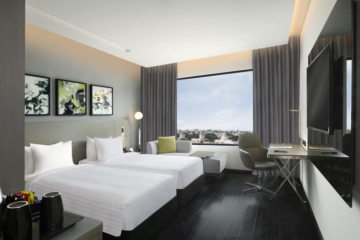 Ein modernes Hotelzimmer mit zwei Betten, einem Schreibtisch, einem Stuhl, einem Flachbildfernseher und einem großen Fenster mit Blick auf die Stadt.