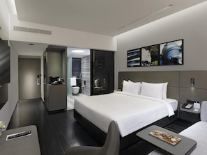 Et moderne hotelværelse med en stor seng, et skrivebord, en stol, et lille bord med dessert, et bruseområde og et fjernsyn.