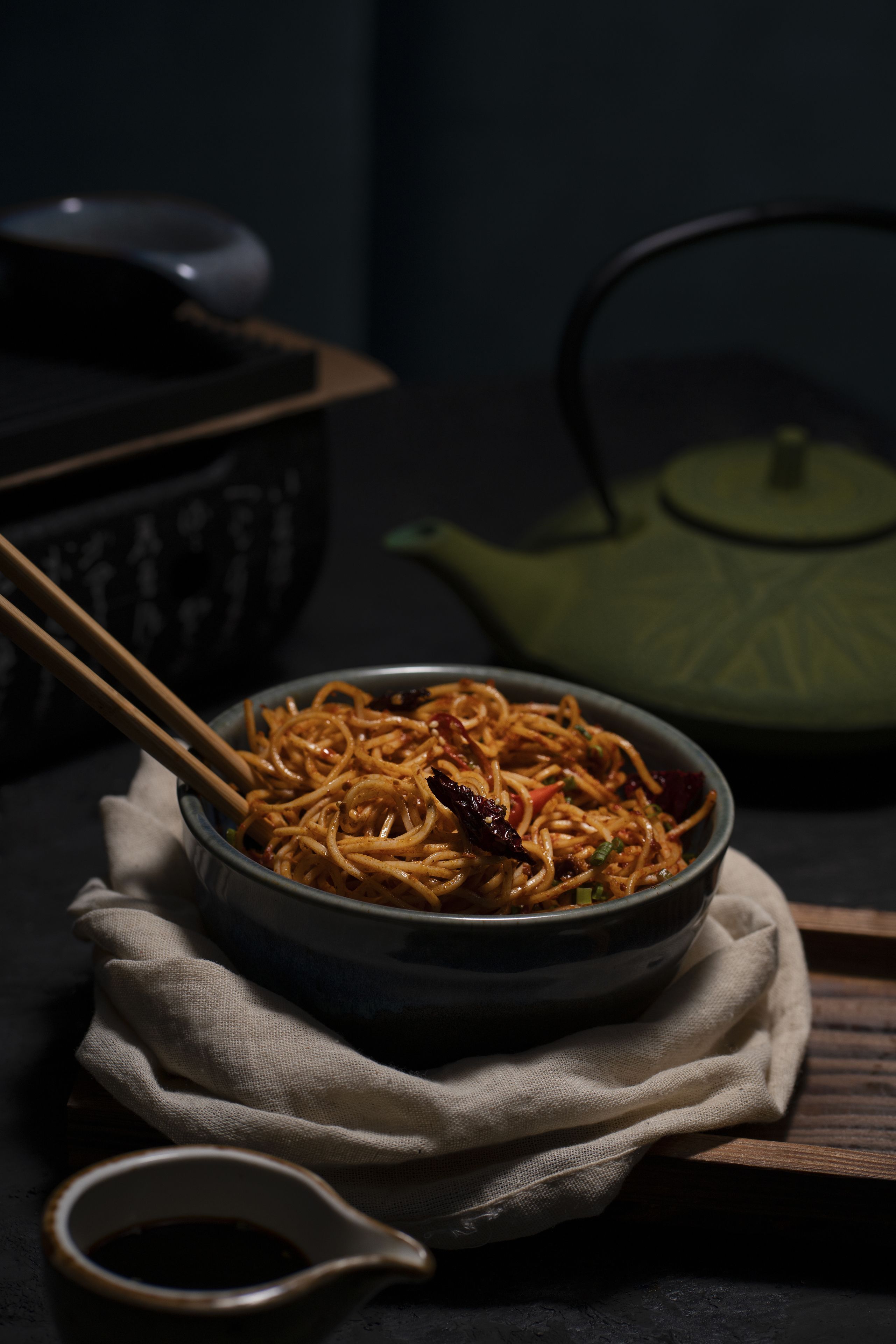 A bowl of noodles with chopsticks, a teapot, and a small bowl of sauce on a table.