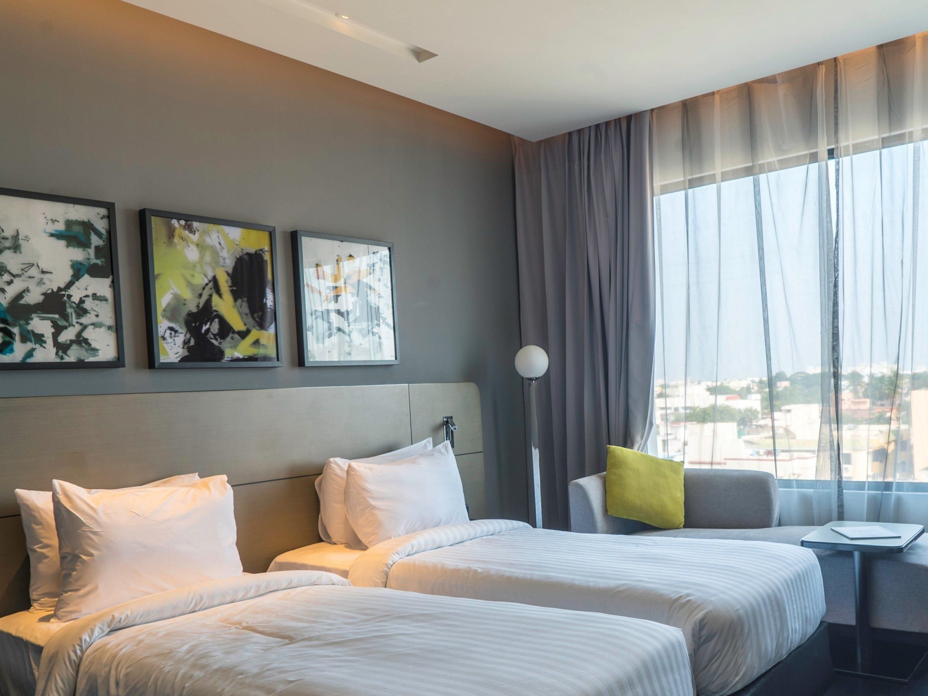 A hotel room with two beds, a nightstand, a lamp, and a window with curtains.