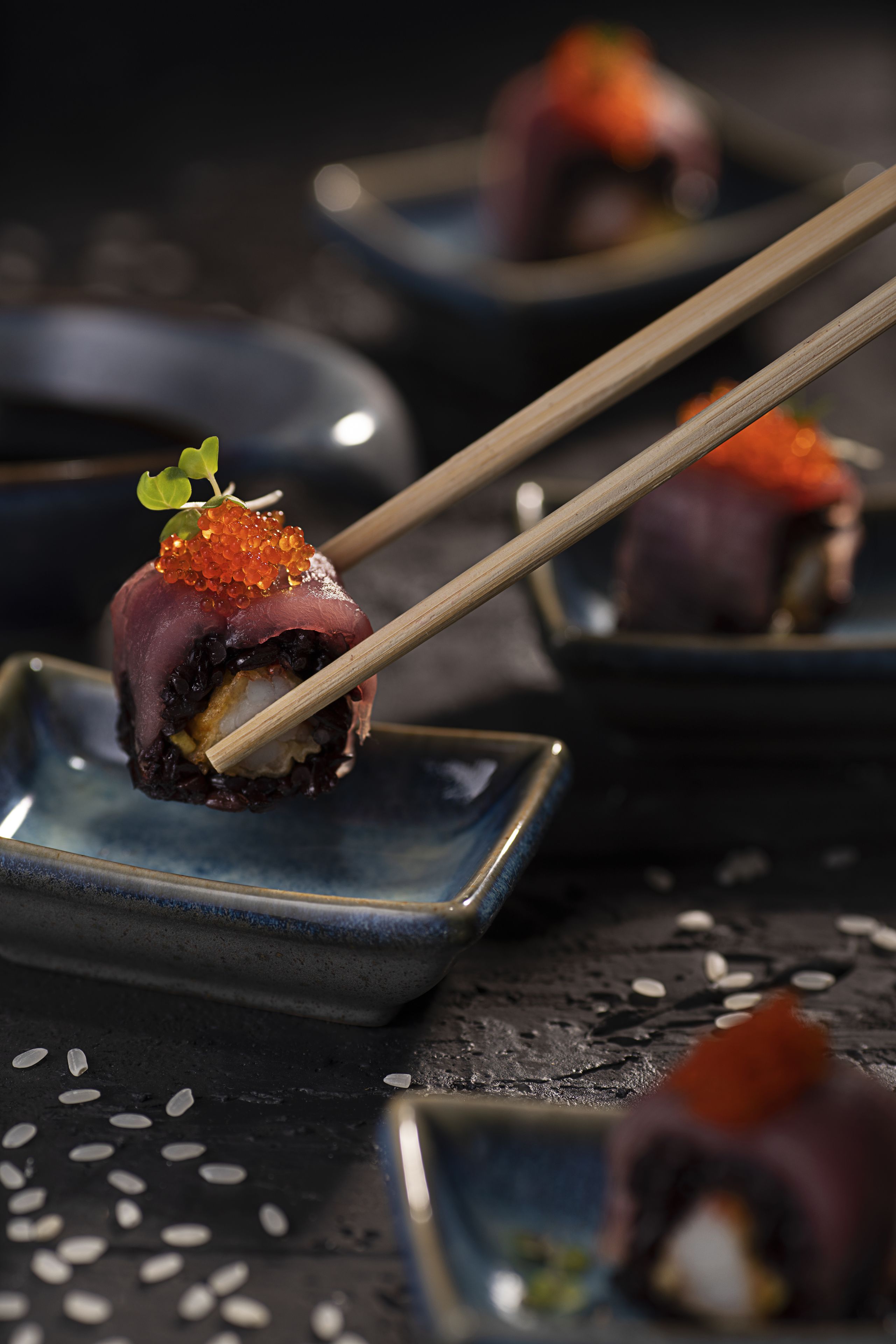 A close-up of sushi with chopsticks, garnished with roe and a leaf, on a dark plate.