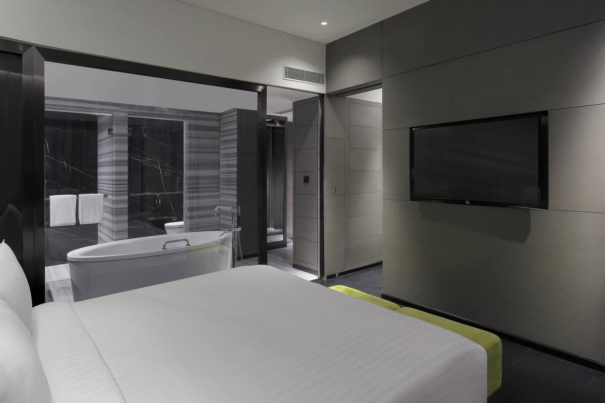 A modern bedroom with a large bed, a TV mounted on the wall, and a bathroom with a bathtub and shower.