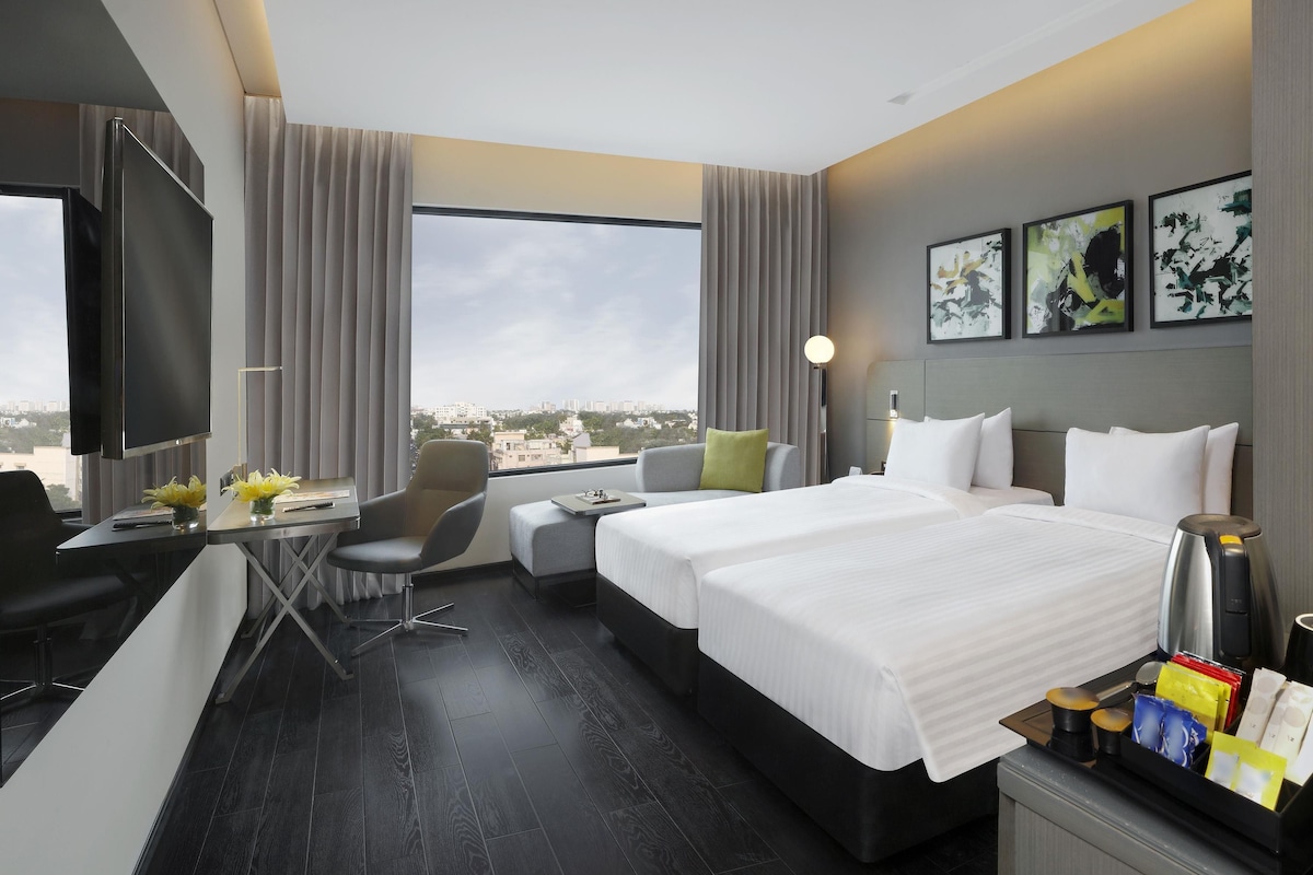 A hotel room with a bed, desk, chair, and a large window offering a city view.
