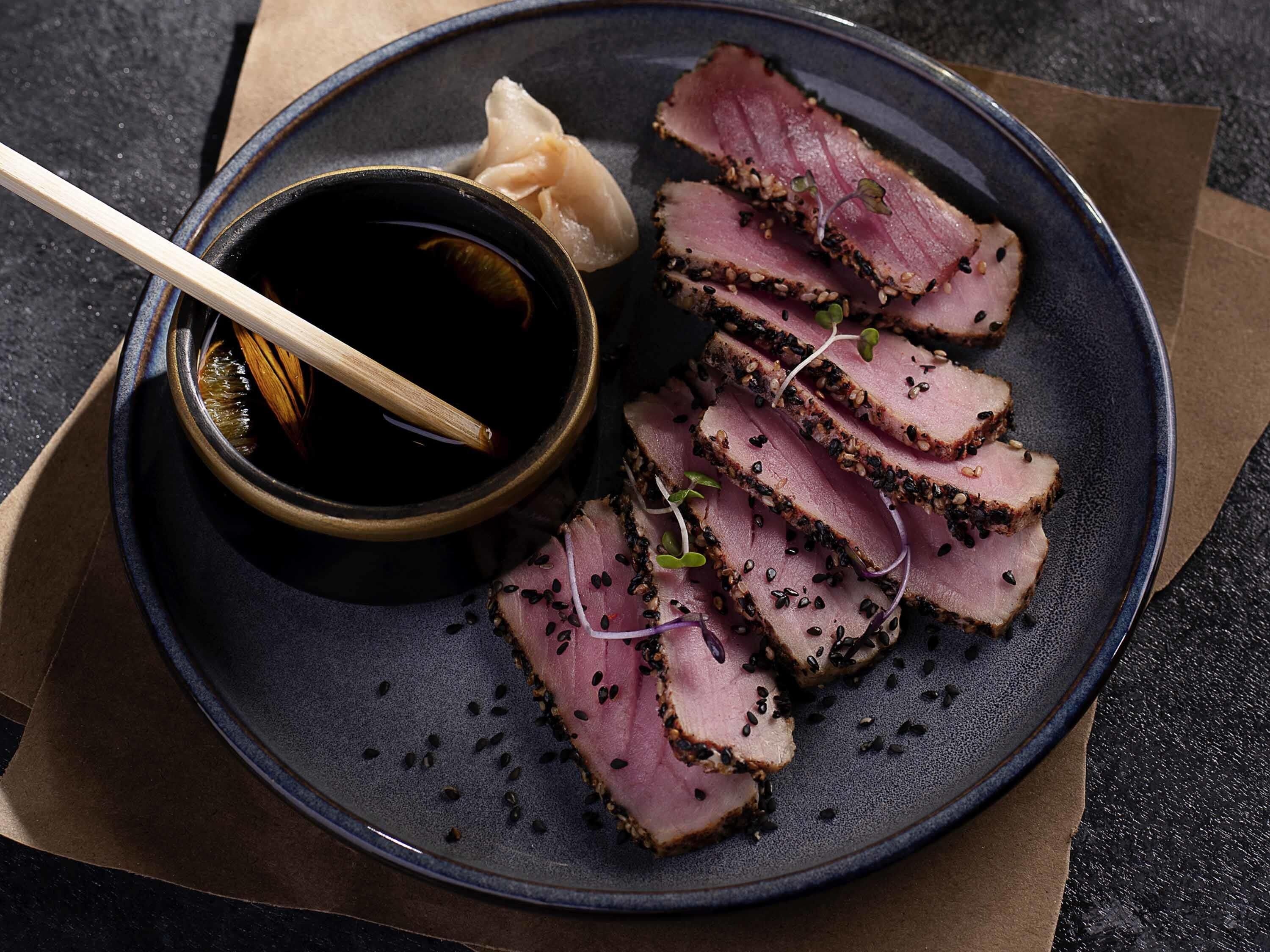 Un plato de carne en rodajas con semillas de sésamo, un tazón de salsa y palillos.