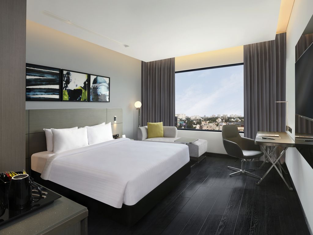 A modern hotel room with a large bed, a desk, a chair, and a view of the city.