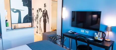 A hotel room with a bed, a desk with a chair, a TV, and a lamp.